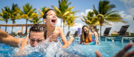 Ein Schwimmbecken mit Personen im Wasser, Palmen und Liegestühlen im Hintergrund.