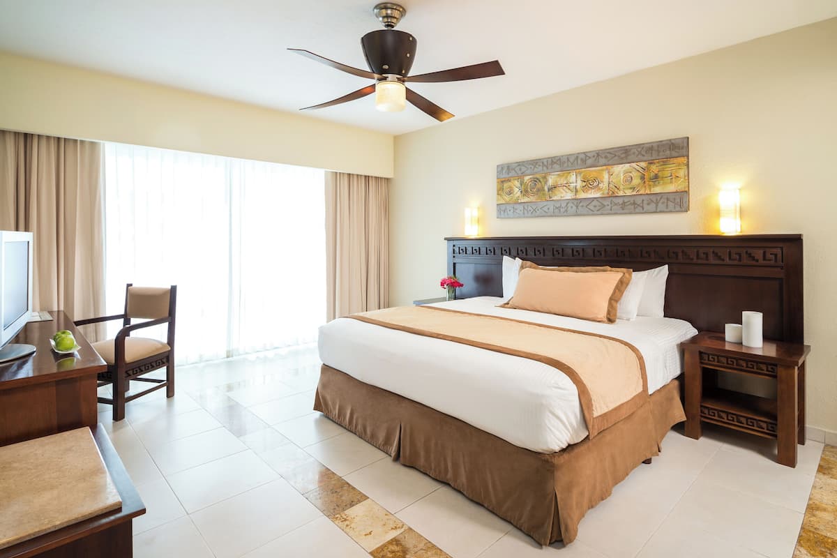 Habitación de hotel con una cama grande, un escritorio con silla, un ventilador de techo y una obra de arte colgada en la pared.