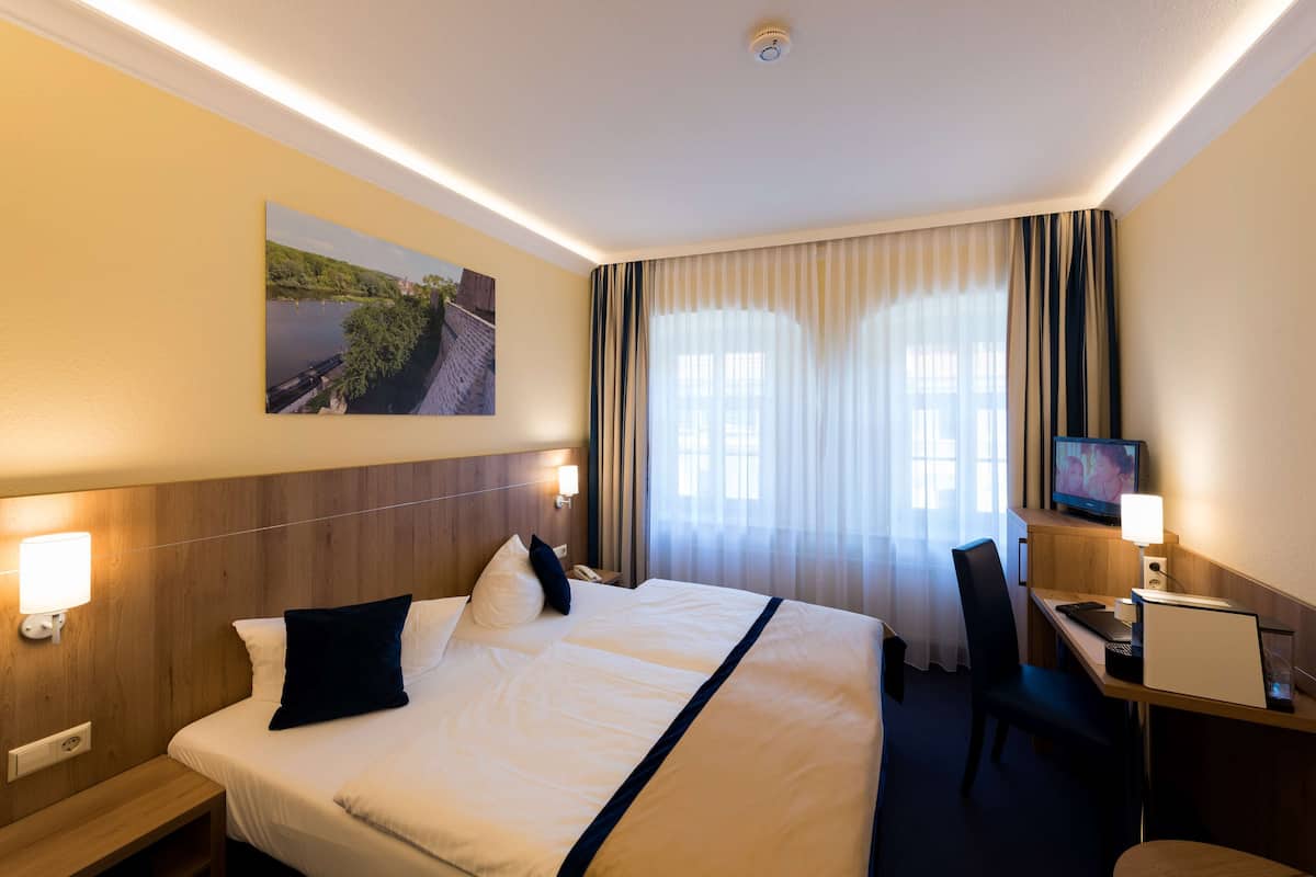 A hotel room with a bed, a desk with a computer, a television, and a painting on the wall.
