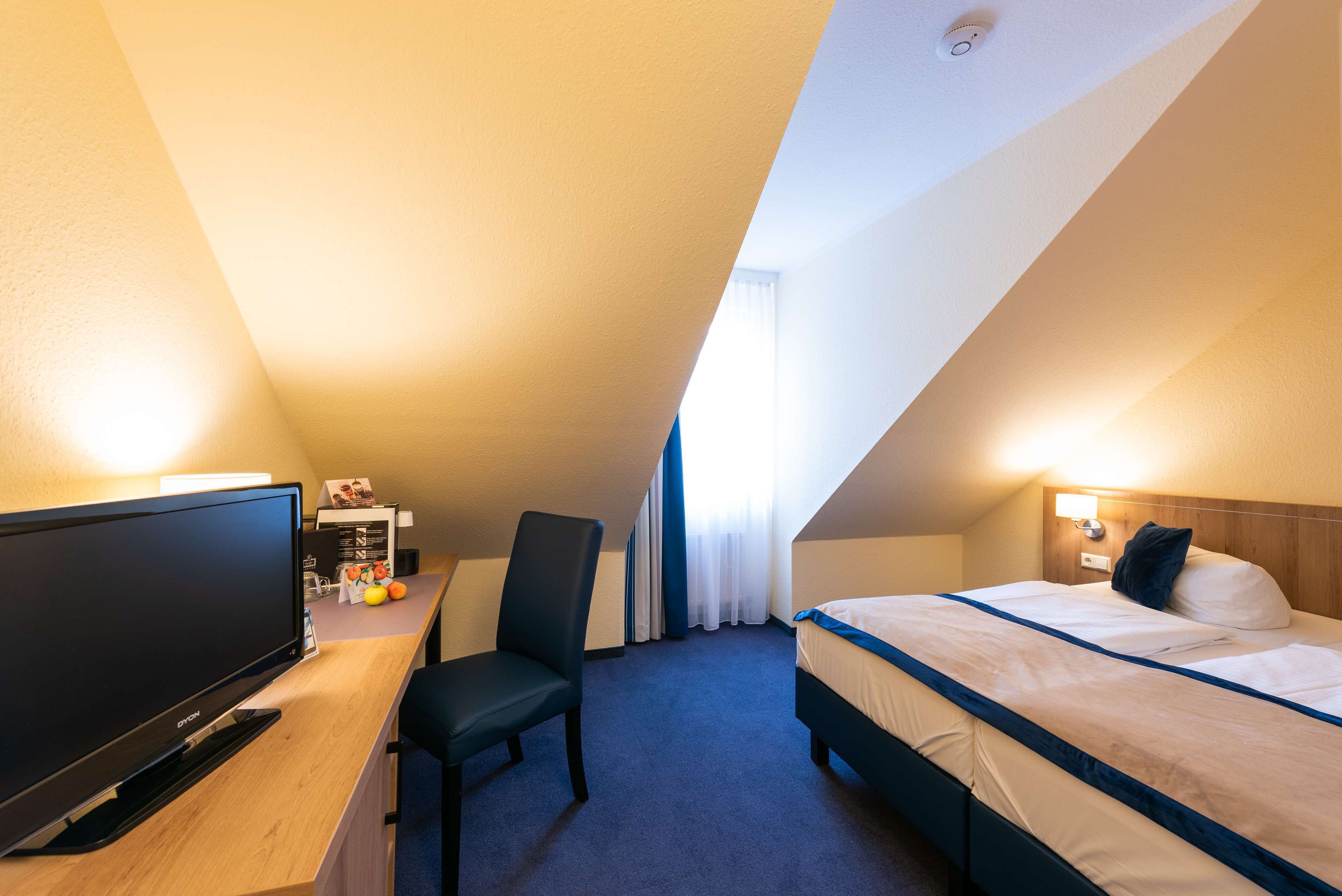 A hotel room with a flat-screen TV, a desk, a chair, a bed with a headboard, and a window with curtains.