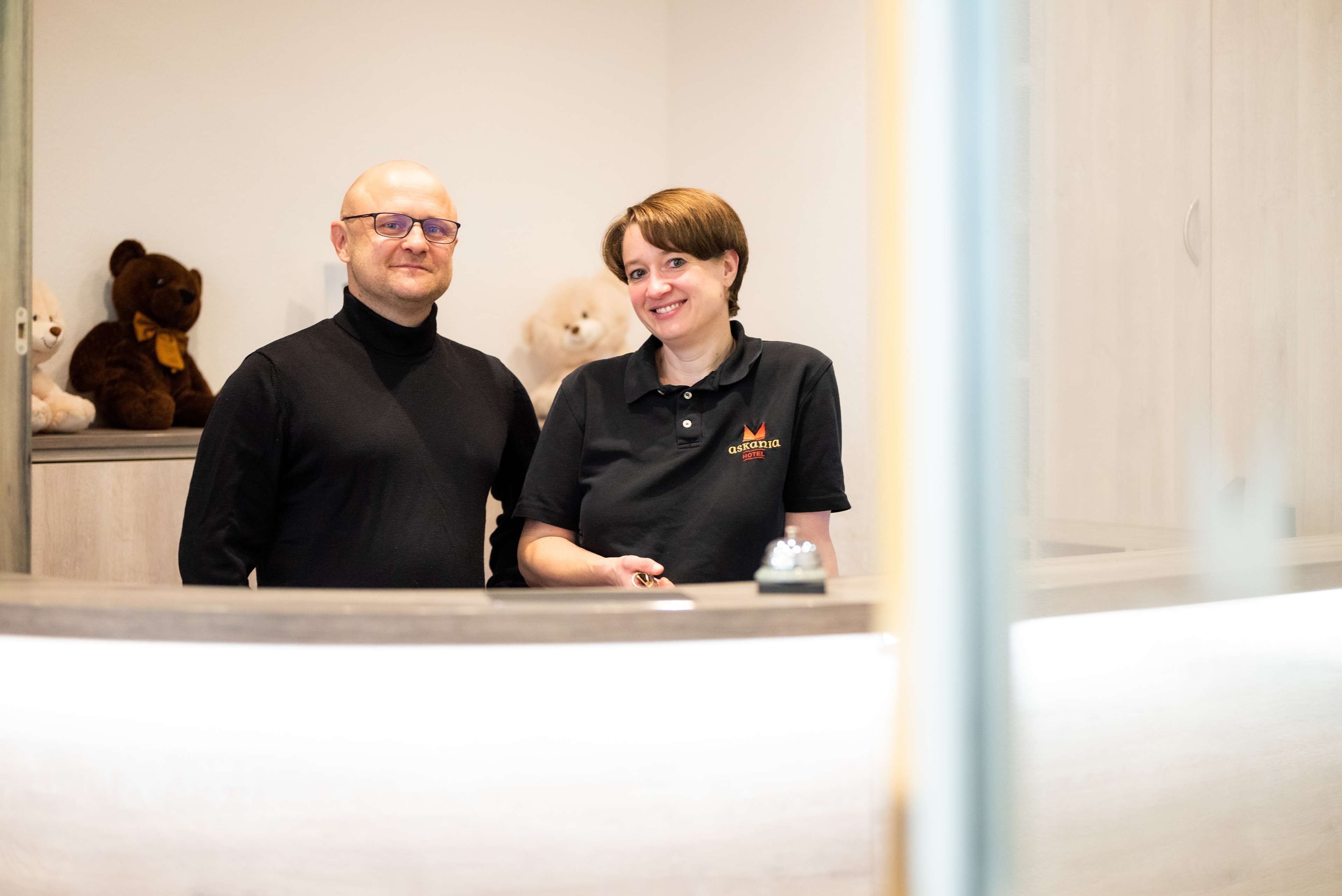 Deux personnes se tiennent dans le hall d’un hôtel, près d’un comptoir d’accueil, avec un ours en peluche et un petit chien.