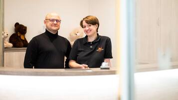 Deux personnes se tiennent dans le hall d’un hôtel, près d’un comptoir d’accueil, avec un ours en peluche et un petit chien.