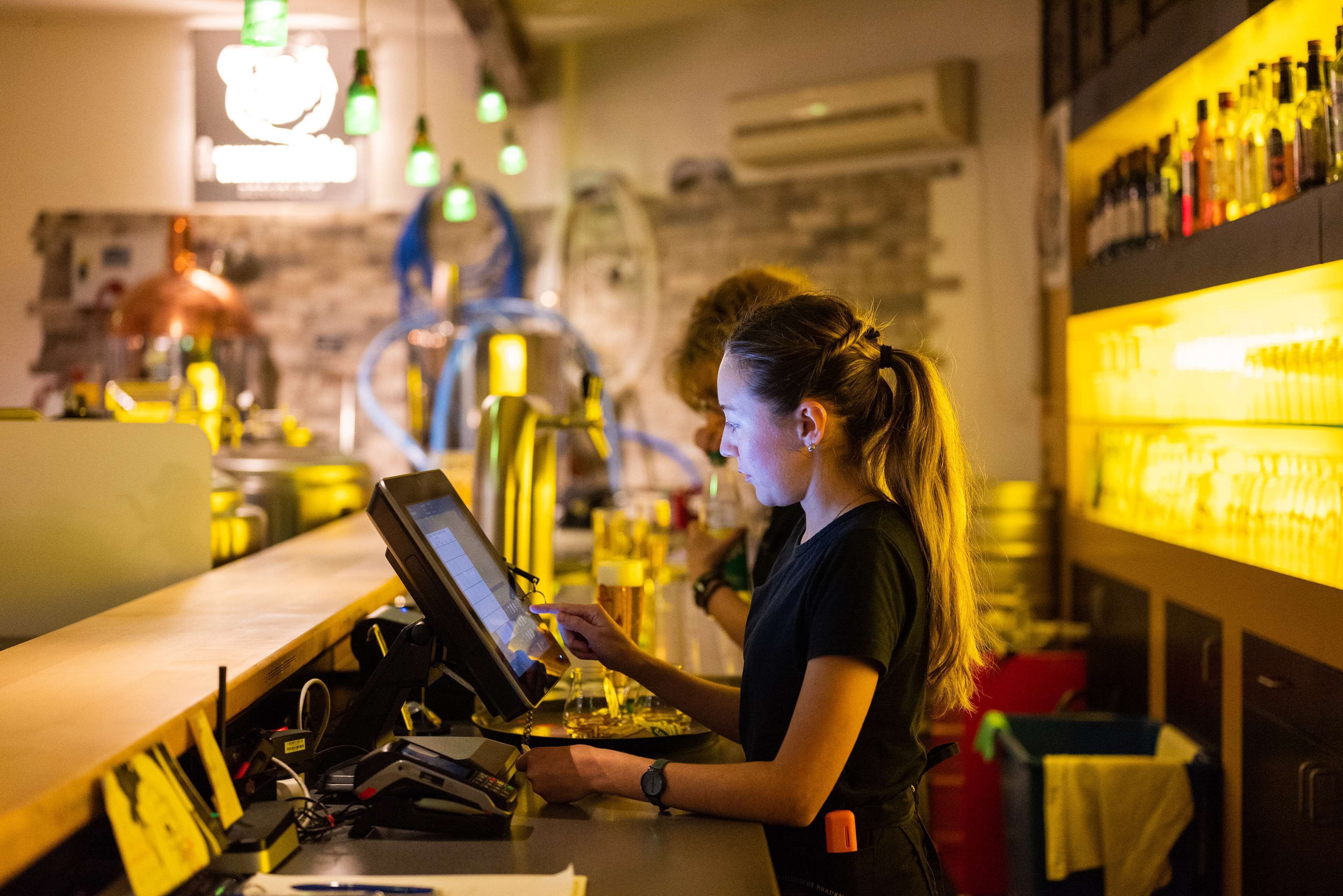 A barista at work in a cafe with a coffee machine, counter, and shelves of bottles.