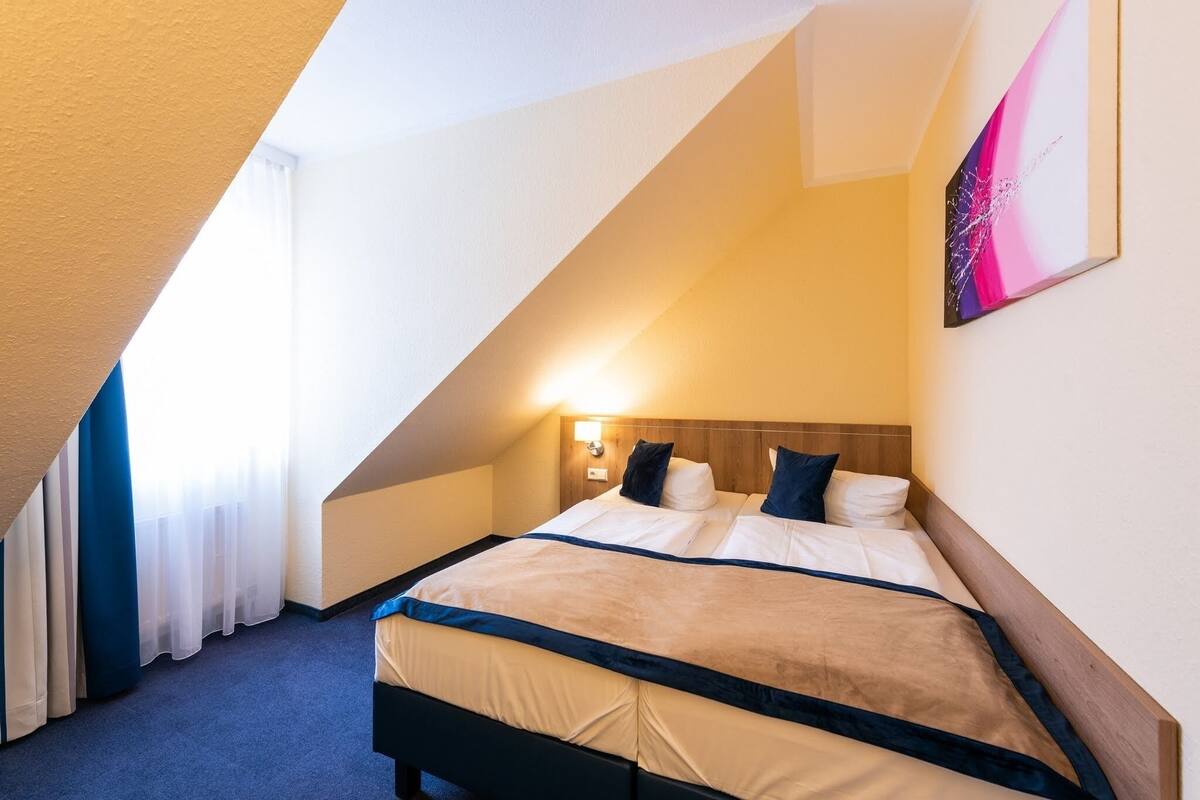 A hotel room with a sloped ceiling, a bed with a wooden headboard, and a wall-mounted light fixture.