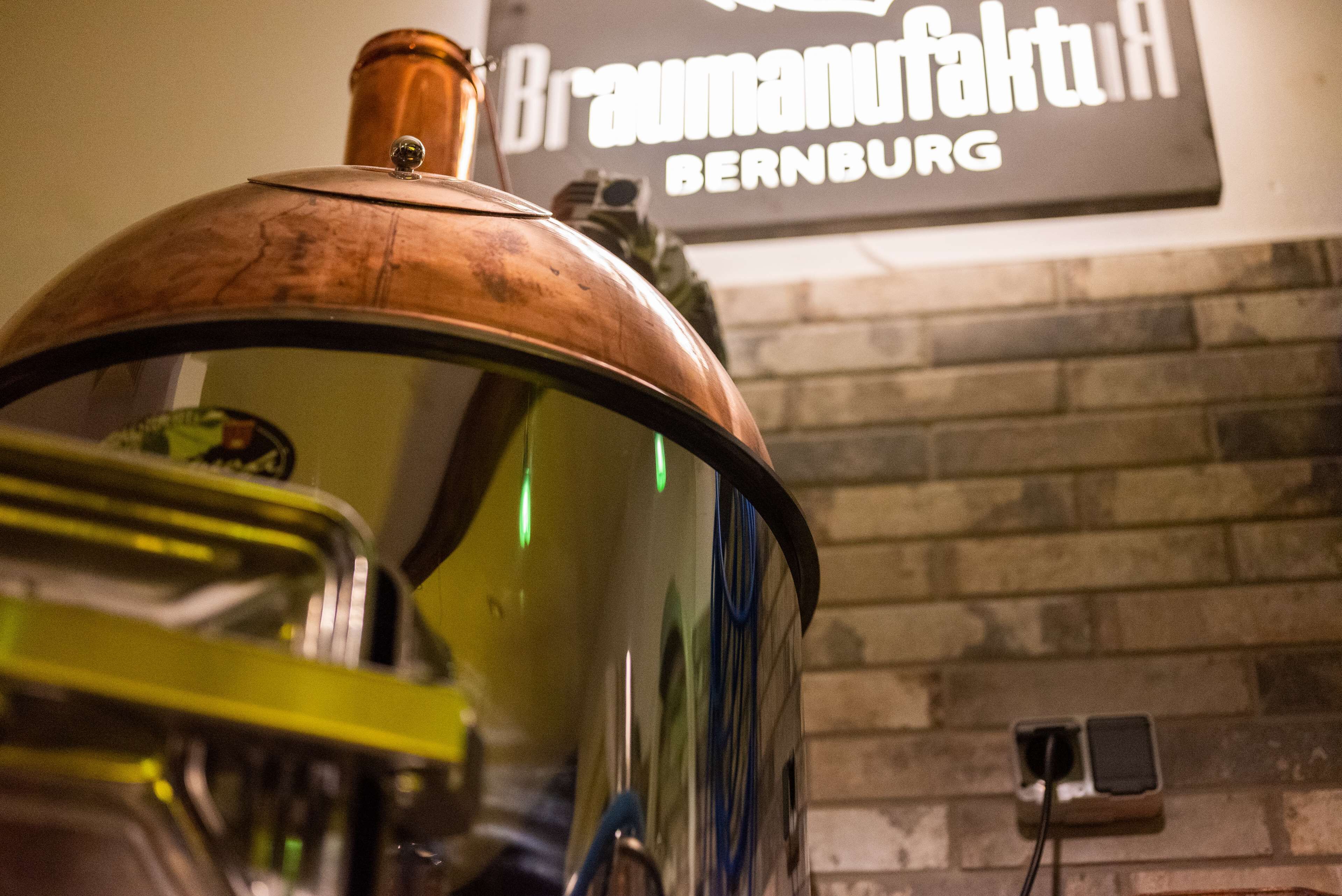A brass-colored object with a reflective surface, possibly a part of a vehicle or machinery, in front of a brick wall with a sign that reads "Braumanufaktur BERNBURG".