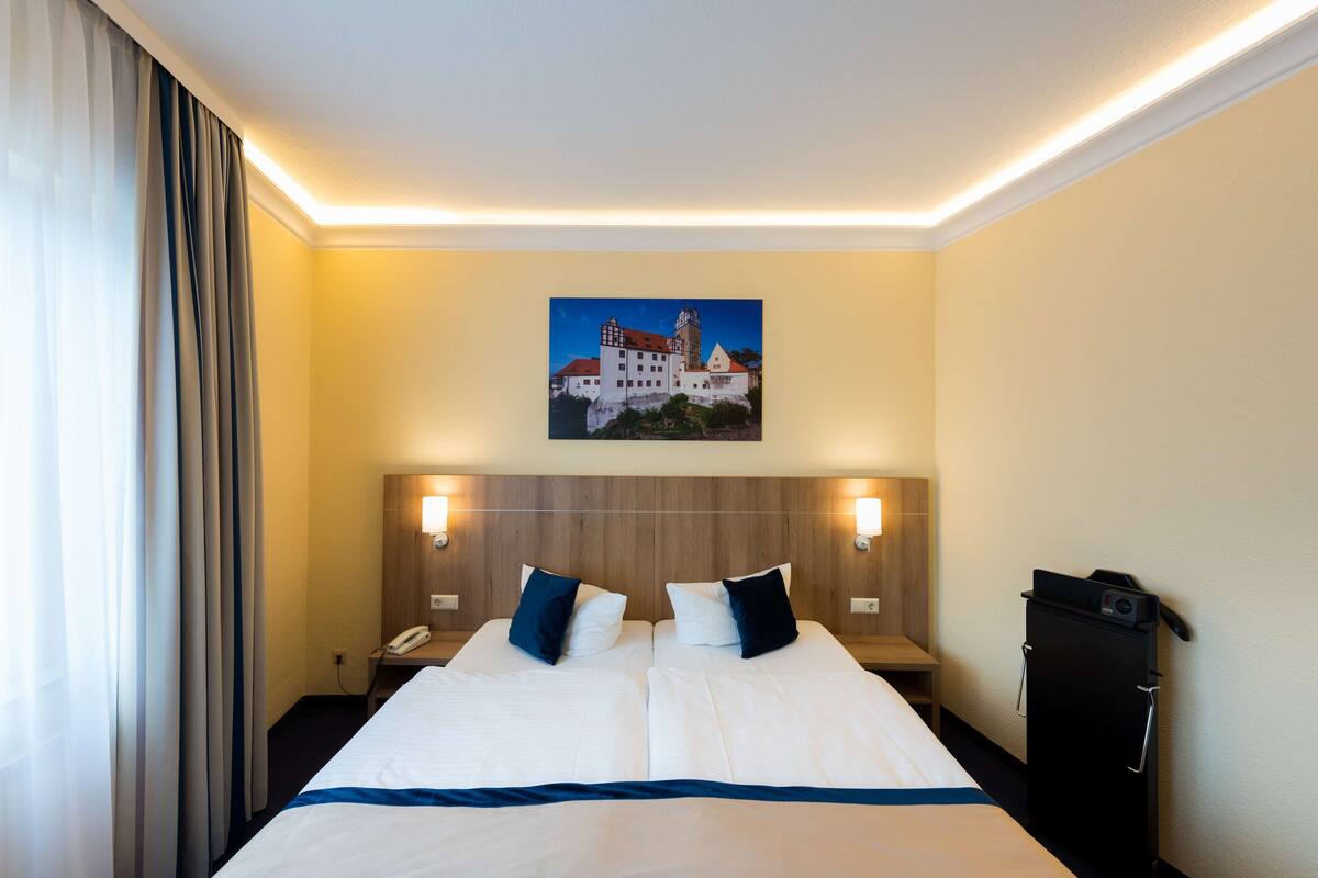 A hotel room with a large bed, two bedside tables, a nightstand, a wall-mounted painting, and a black cabinet.