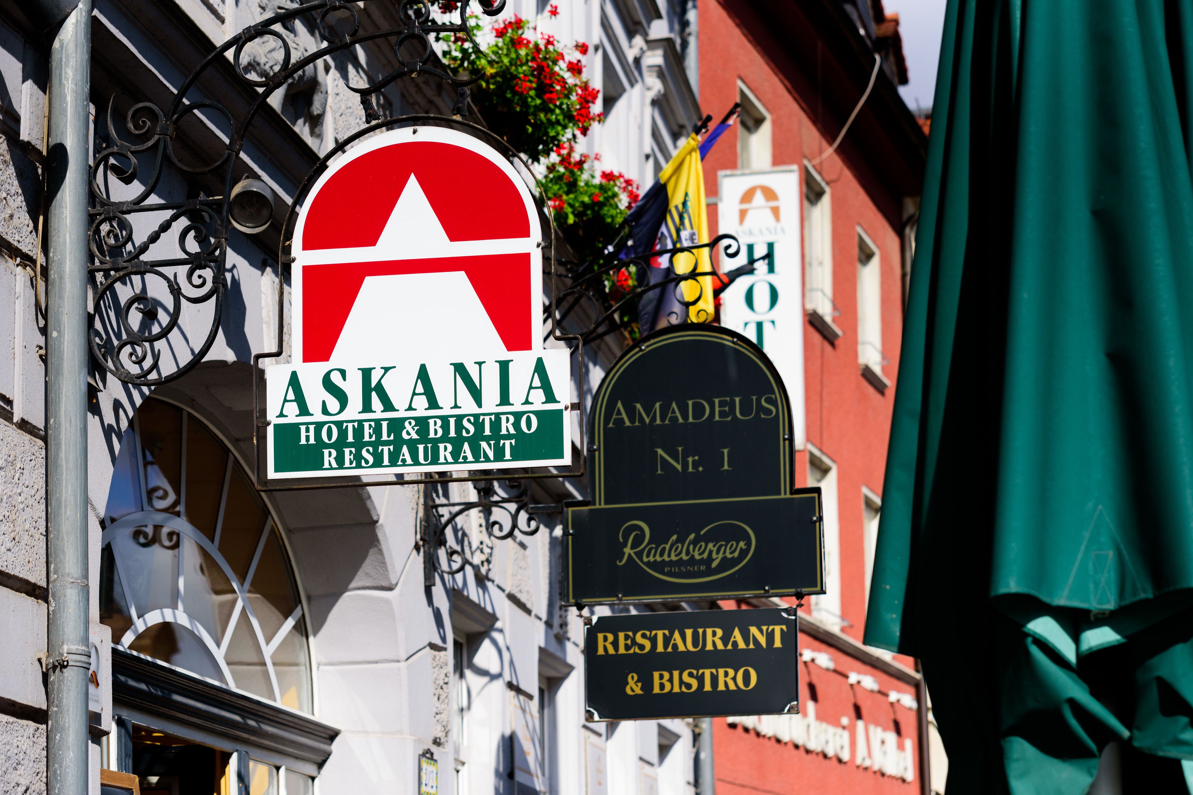 A street sign for Askania Hotel & Bistro Restaurant, with a red and white logo, and adjacent signs for Amadeus Hotel Nr. 1 and Radeberger Pilsner.