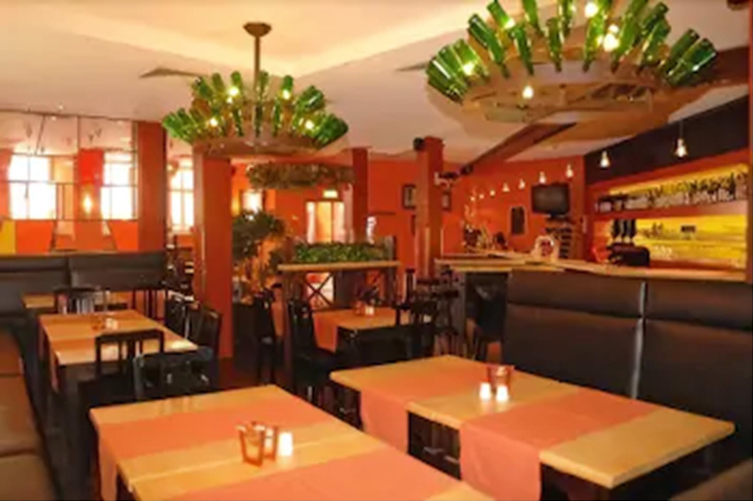 Interior de un restaurante con cabinas y mesas, una zona de bar y un techo decorado con botellas verdes colgantes.