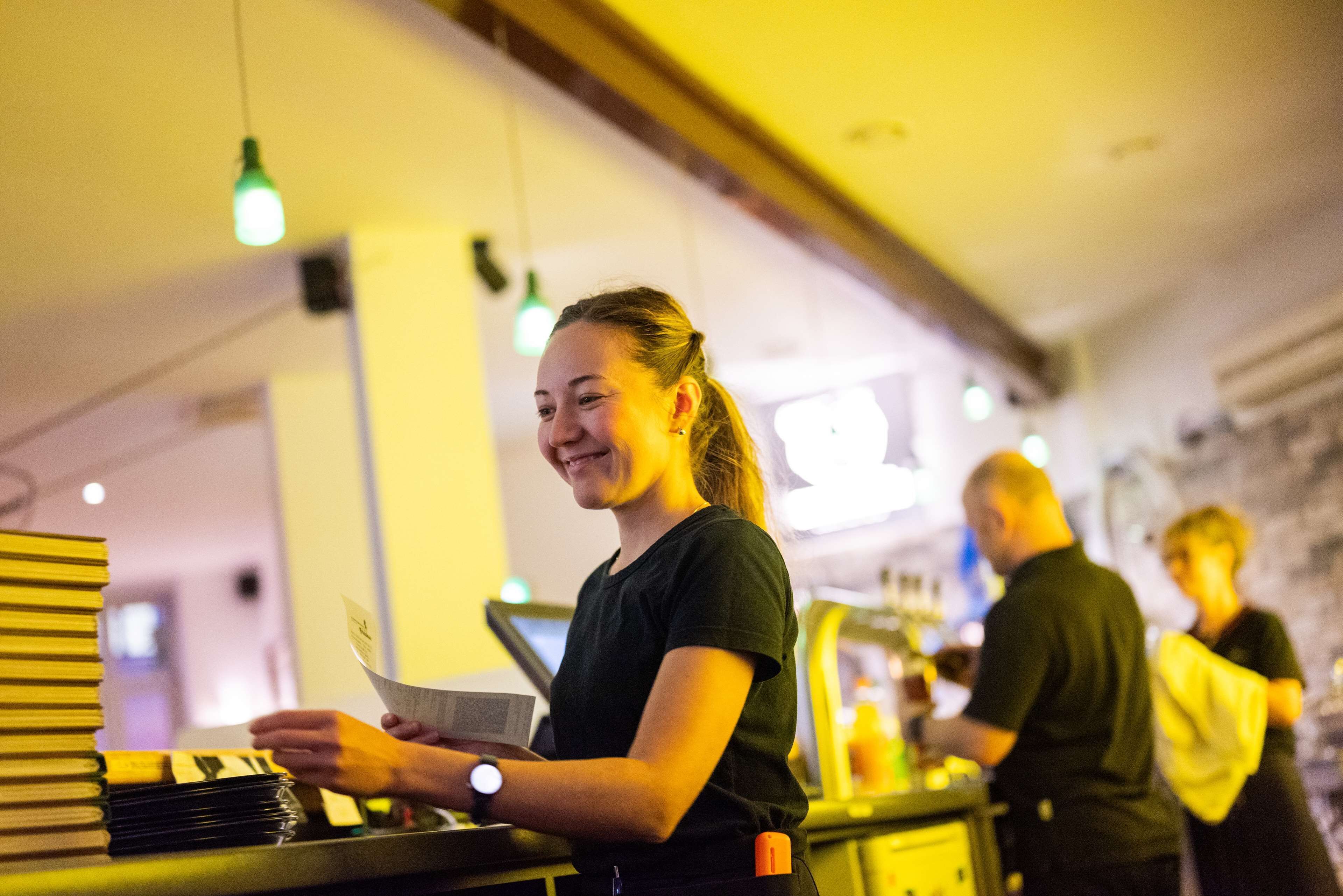 A bar setting with a person holding a menu, another person working behind the bar, and a stack of books on the counter.