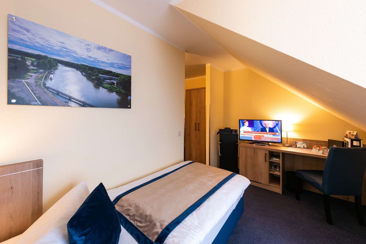 A hotel room with a bed, a desk with a computer, a TV, and a painting of a river.