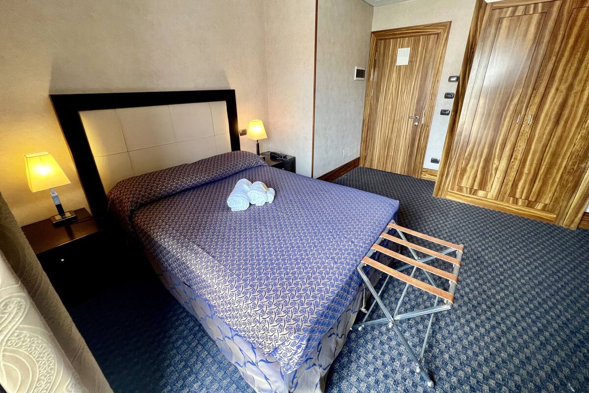 Une chambre d’hôtel avec un lit, une table de chevet, une lampe, une armoire et une serviette pliée posée sur le lit.