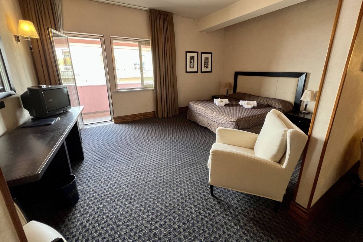 Une chambre d’hôtel avec un lit, une chaise, un bureau avec une télévision et une fenêtre avec des rideaux.