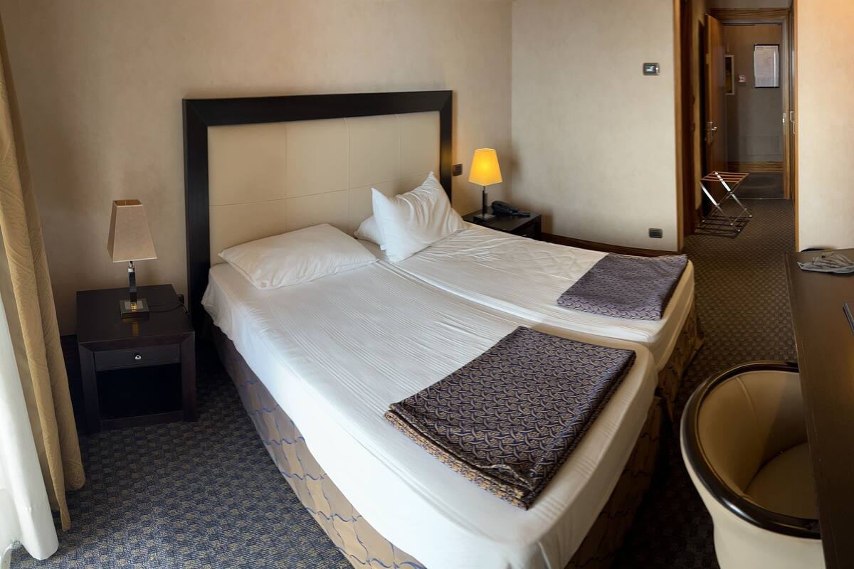 Une chambre d’hôtel avec deux lits, une table de chevet avec une lampe, un bureau avec une télécommande et une salle de bain visible à travers une porte ouverte.