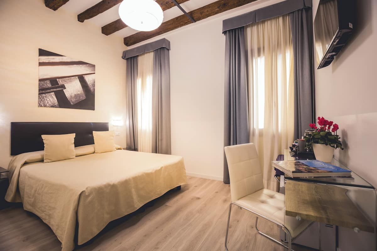 A hotel room with a bed, a desk, a chair, a potted plant, and a painting on the wall.