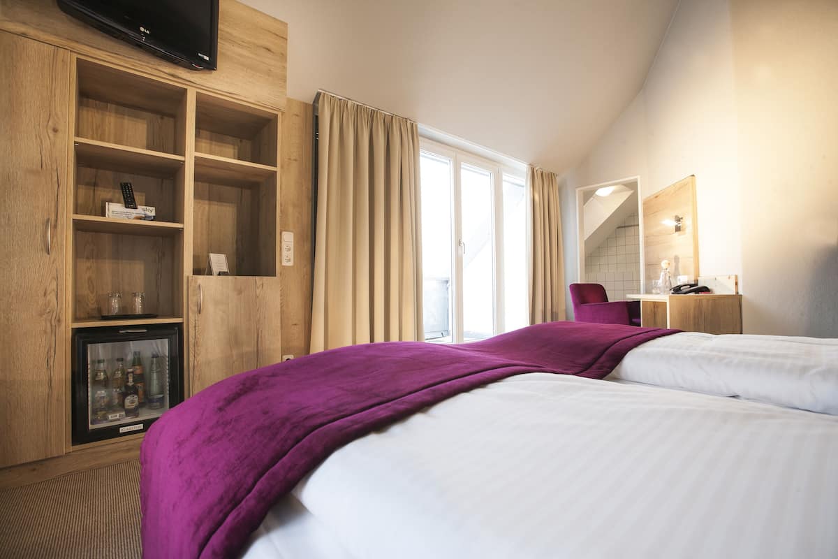Ein Hotelzimmer mit zwei Betten, einer Minibar, einem Fernseher, einem Schreibtisch mit Stuhl und einem großen Fenster mit Vorhängen.