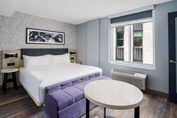 A modern hotel room with a large bed, a round table, a small stool, a wall-mounted picture, and a window with blinds. - Manhattan Centre Hotel (New York)