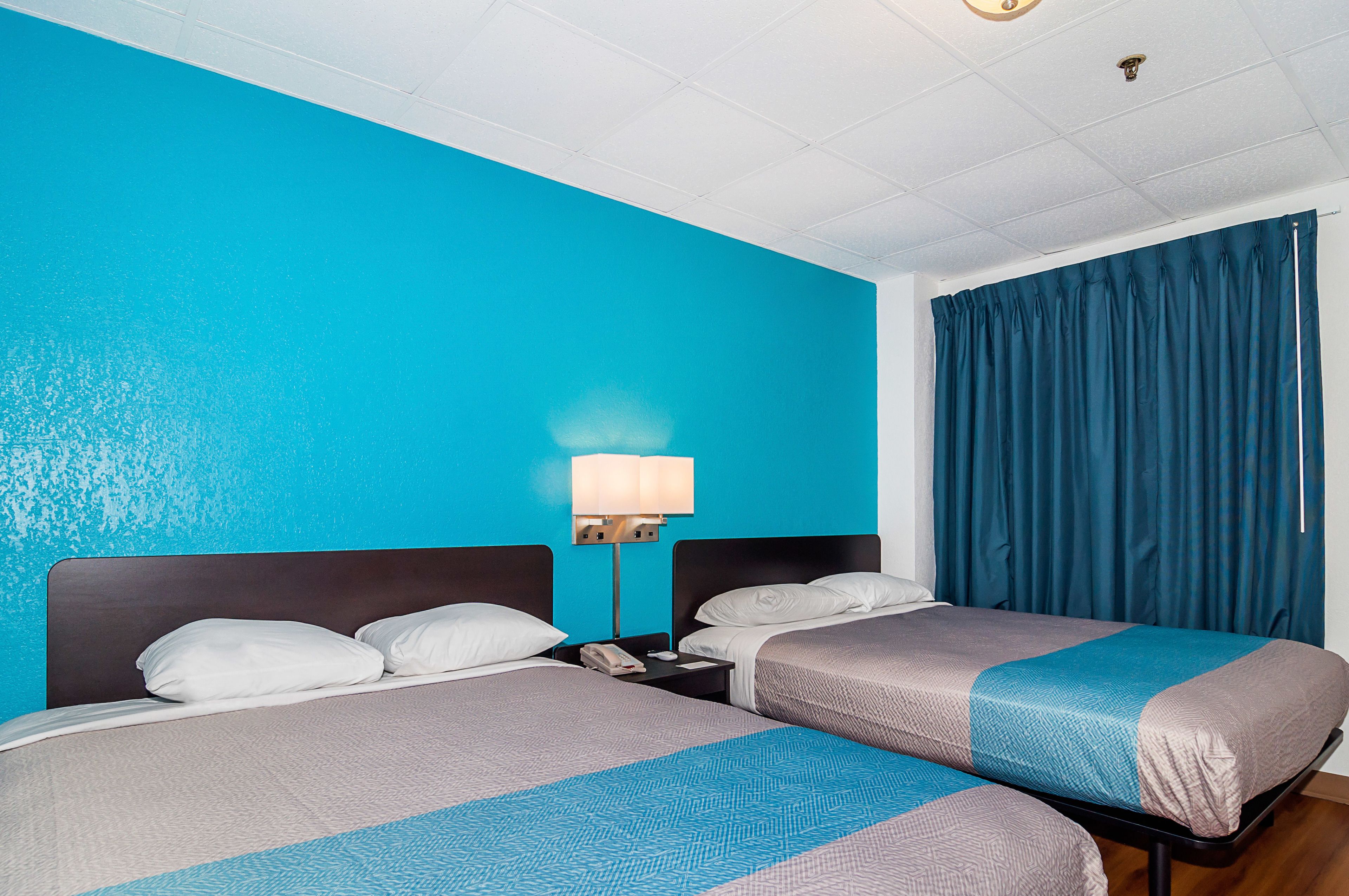 Une chambre d’hôtel avec deux lits, une table de chevet, une lampe et une fenêtre avec des rideaux bleus.