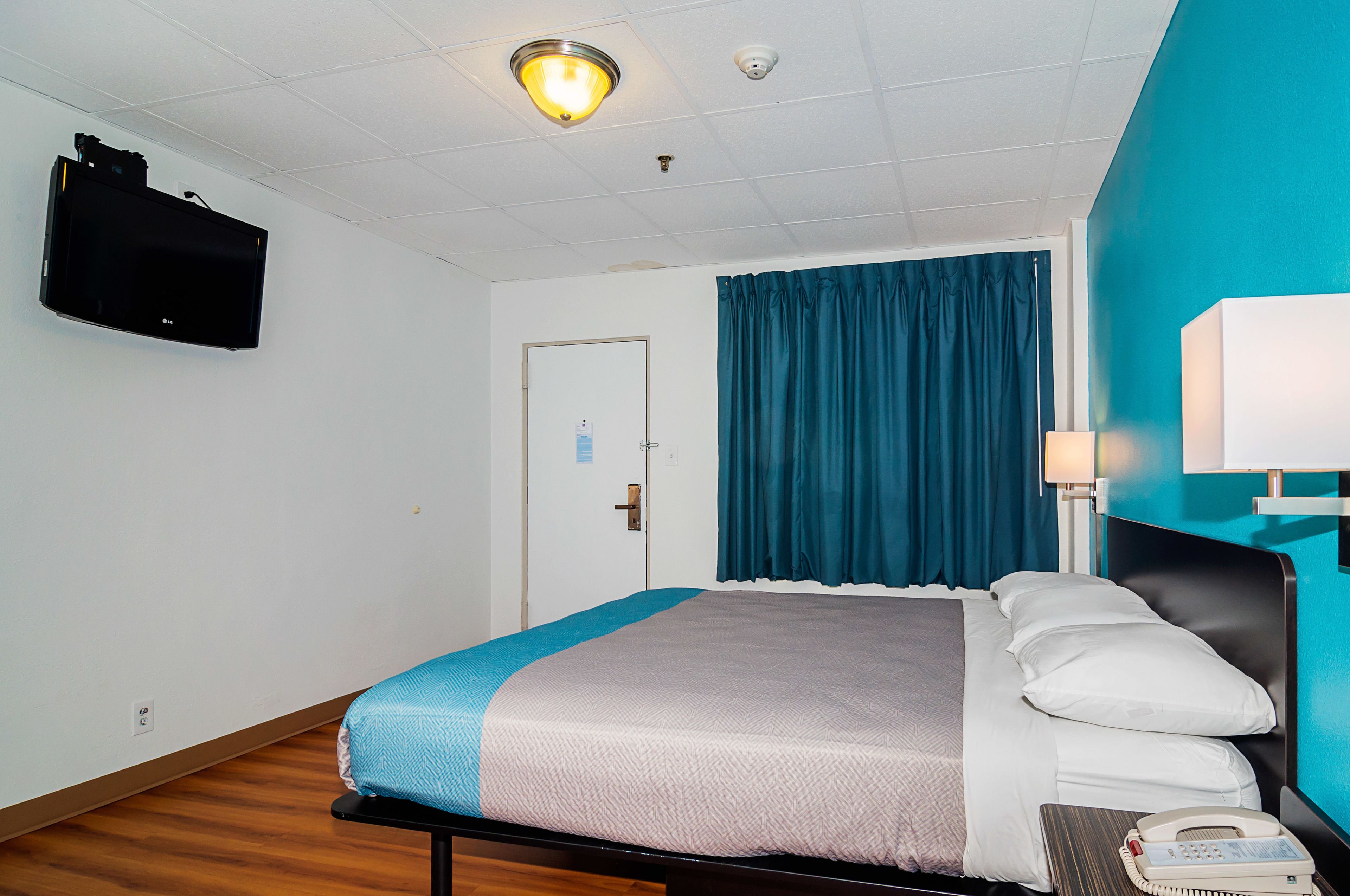Habitación de hotel con cama, escritorio con lámpara, teléfono, televisor montado en la pared y una puerta con un cartel.