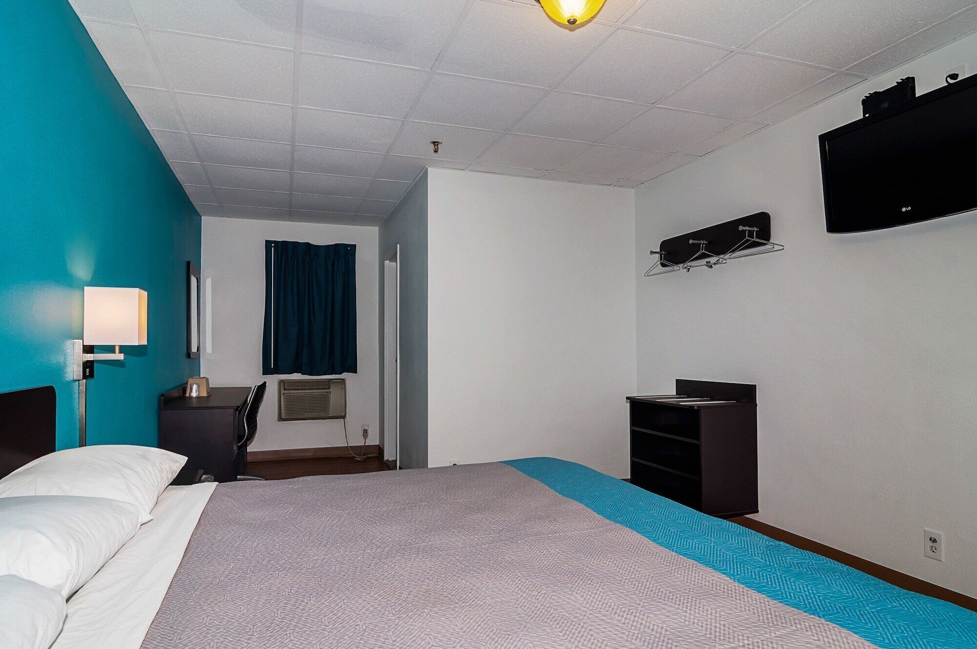 Una camera d'albergo con un letto, una scrivania, una sedia, una lampada da scrivania, una TV e una finestra con tende.