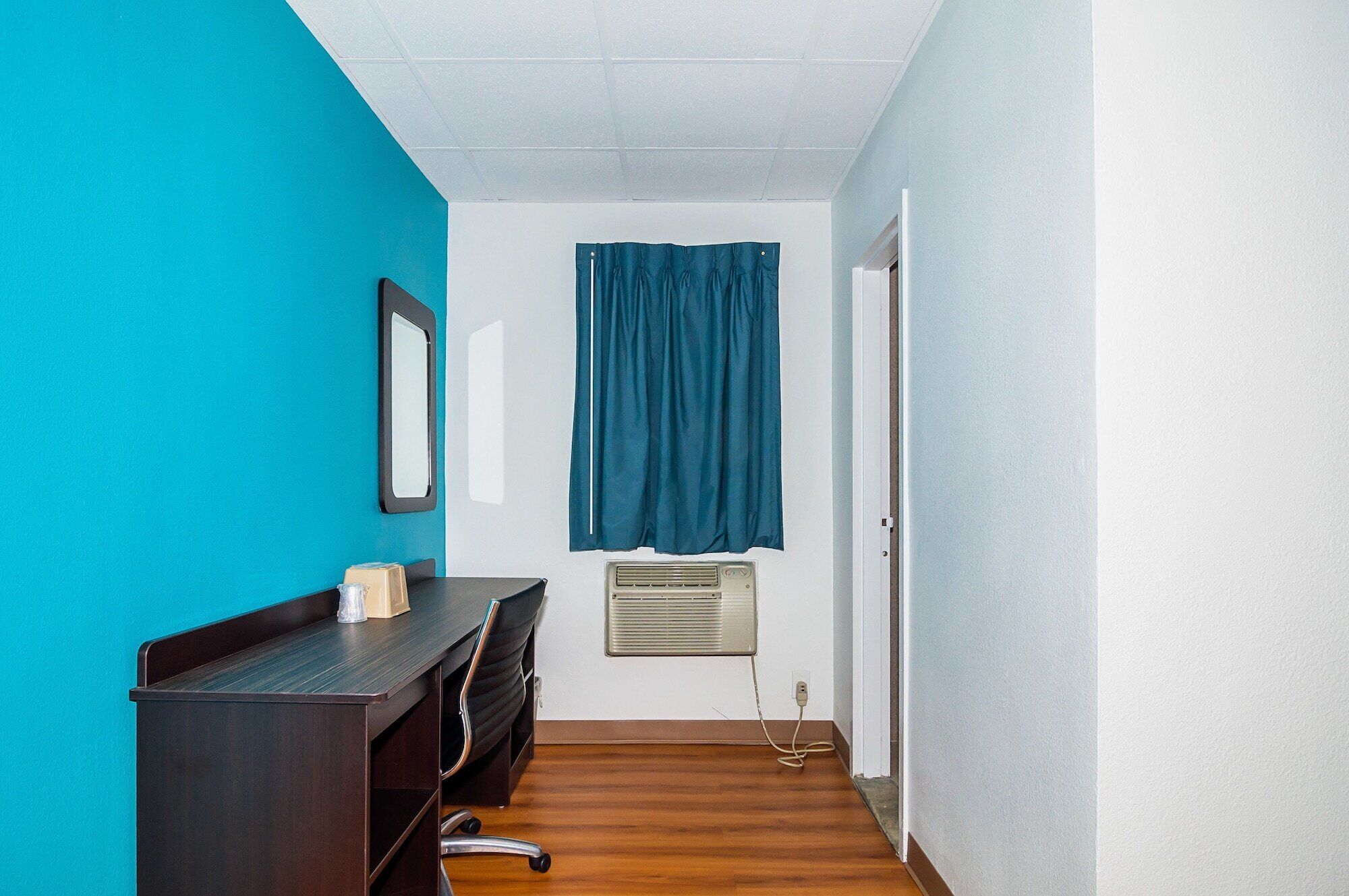 A small, narrow room with a desk, chair, and a window with blue curtains.