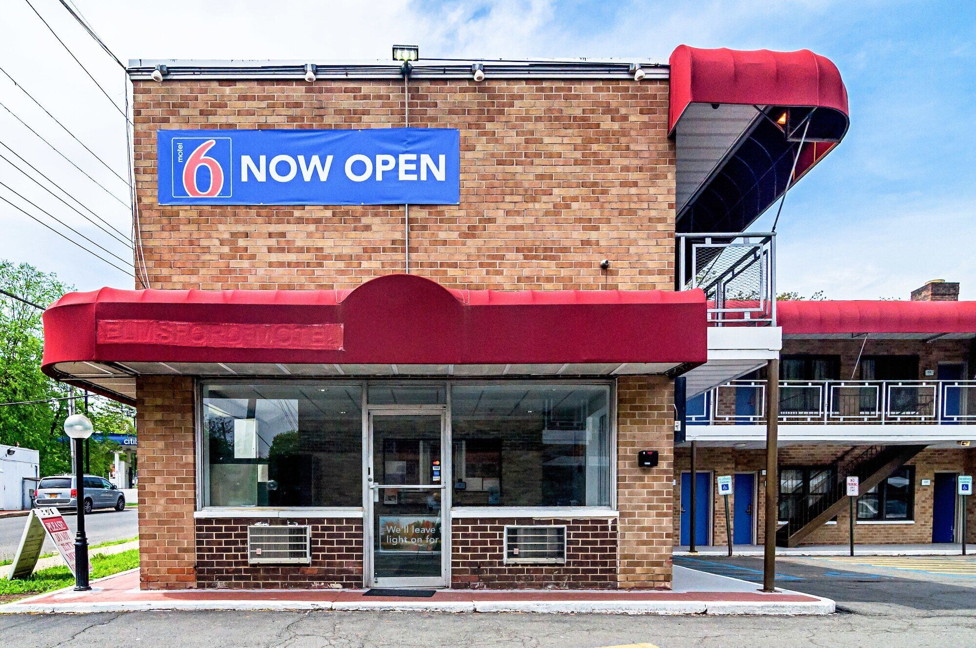 A motel with a '6 Now Open' sign, a brick facade, and a red awning.