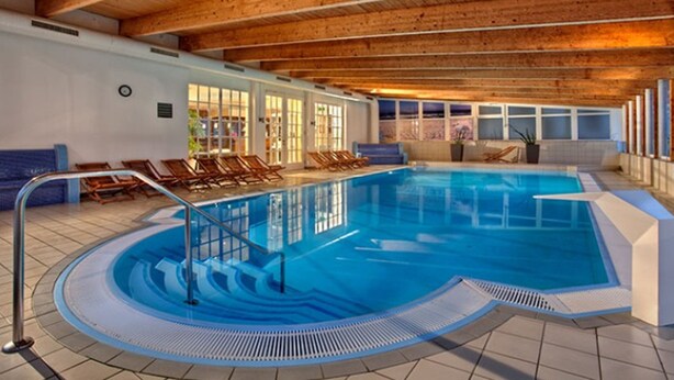 An indoor swimming pool with a wooden ceiling, clear glass walls, and sunlit windows. - Hotel Vier Jahreszeiten Binz  (Binz)