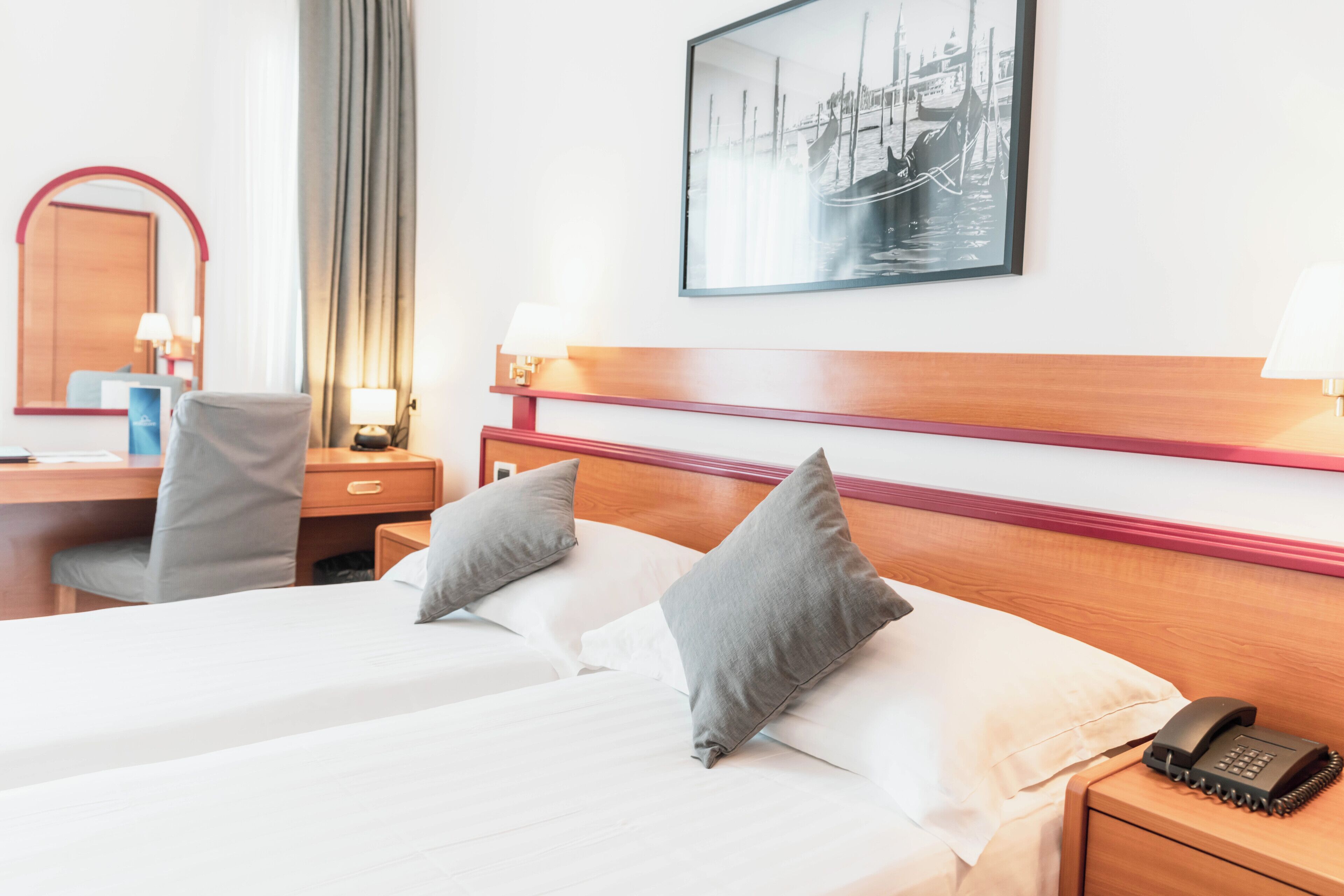 Et hotelværelse med en seng, to puder, et natbord med lampe, et skrivebord med stol, et spejl og et billede i ramme på væggen.