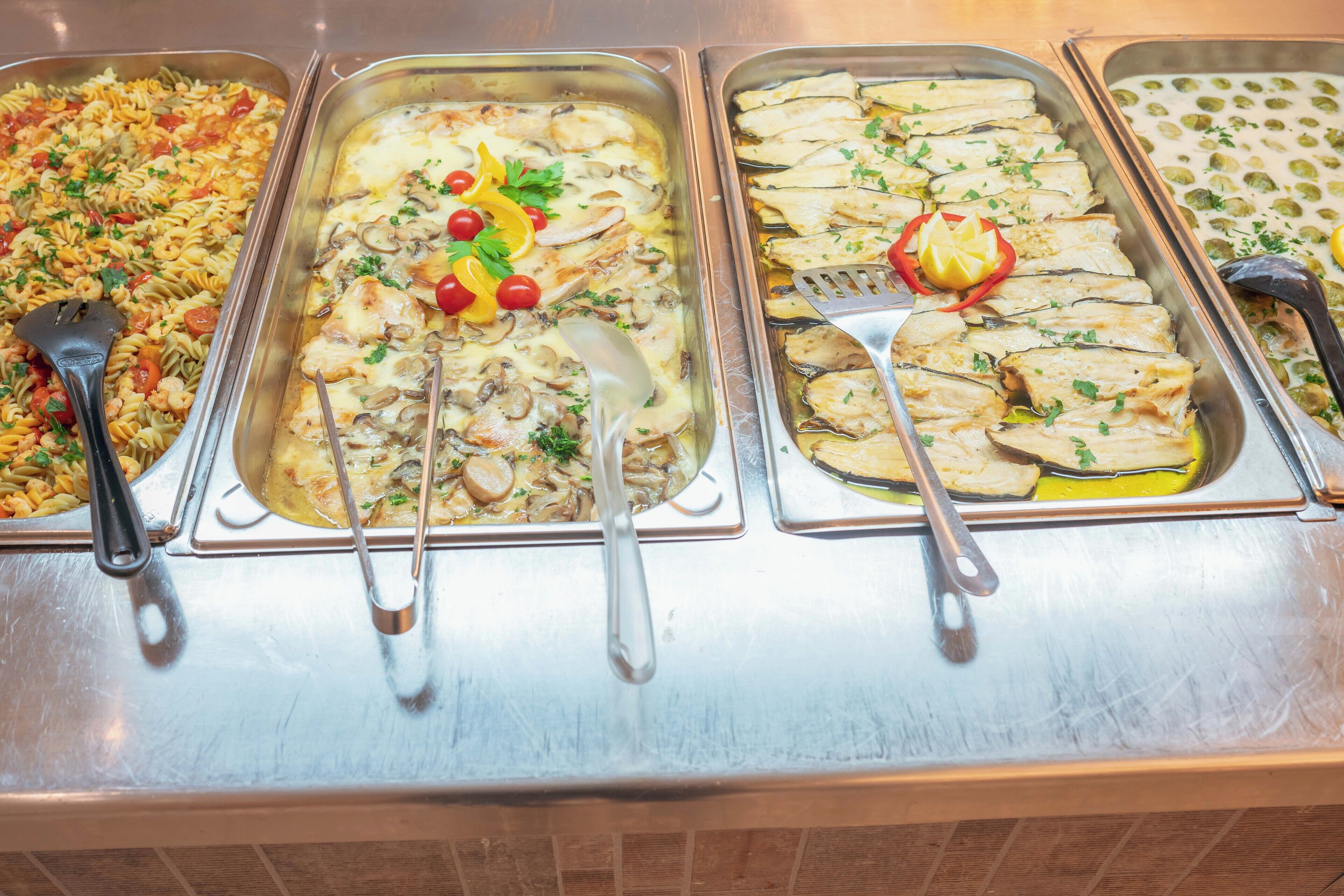 Un bufé con diversos platos, incluyendo pasta, pollo con champiñones, pescado y una ensalada.