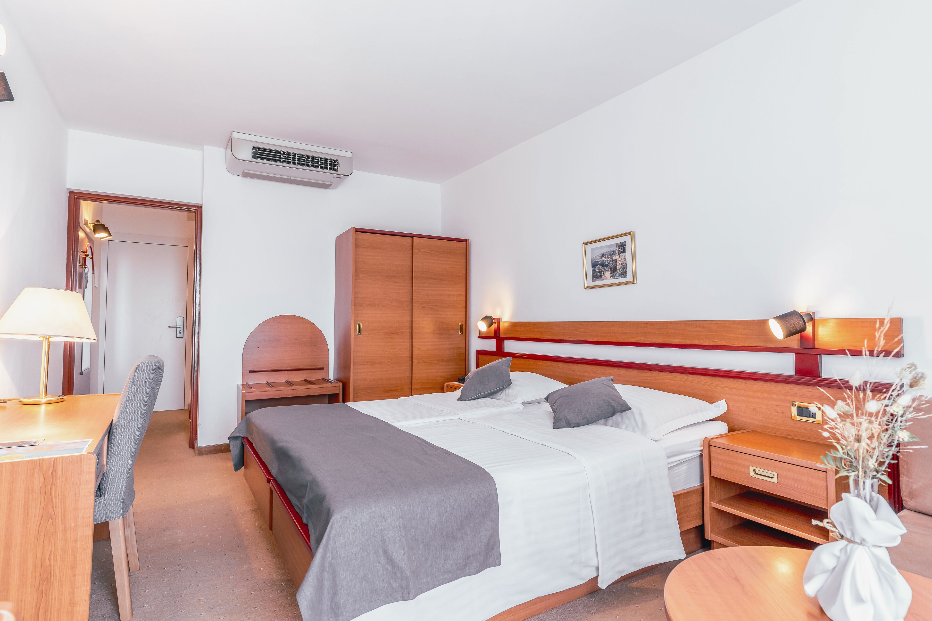 A hotel room with a bed, bedside table, lamp, chair, wardrobe, and an air conditioning unit.