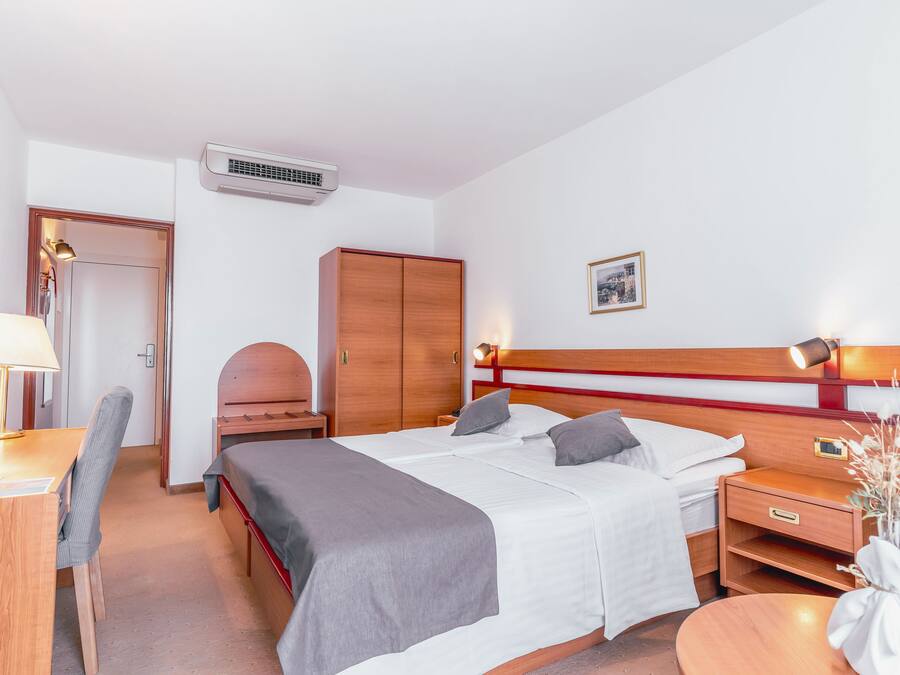 A hotel room with a bed, bedside table, lamp, chair, wardrobe, and an air conditioning unit.