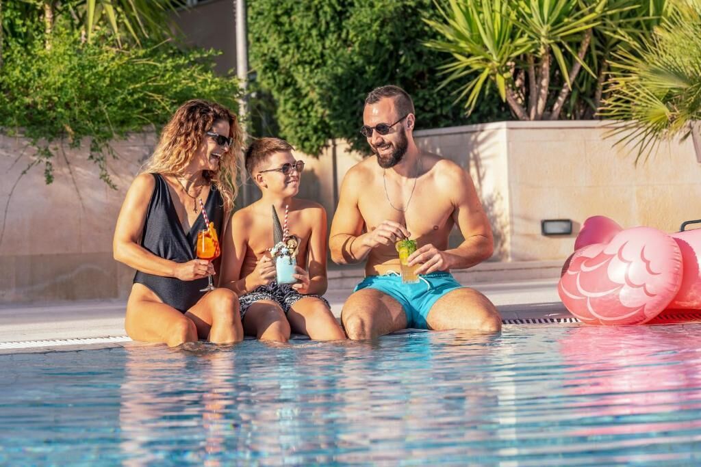Three people sitting by a pool with drinks, surrounded by greenery.