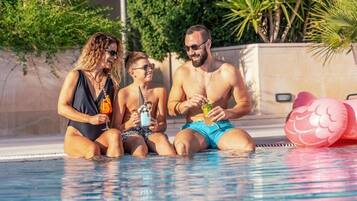 Three people sitting by a pool with drinks, surrounded by greenery.
