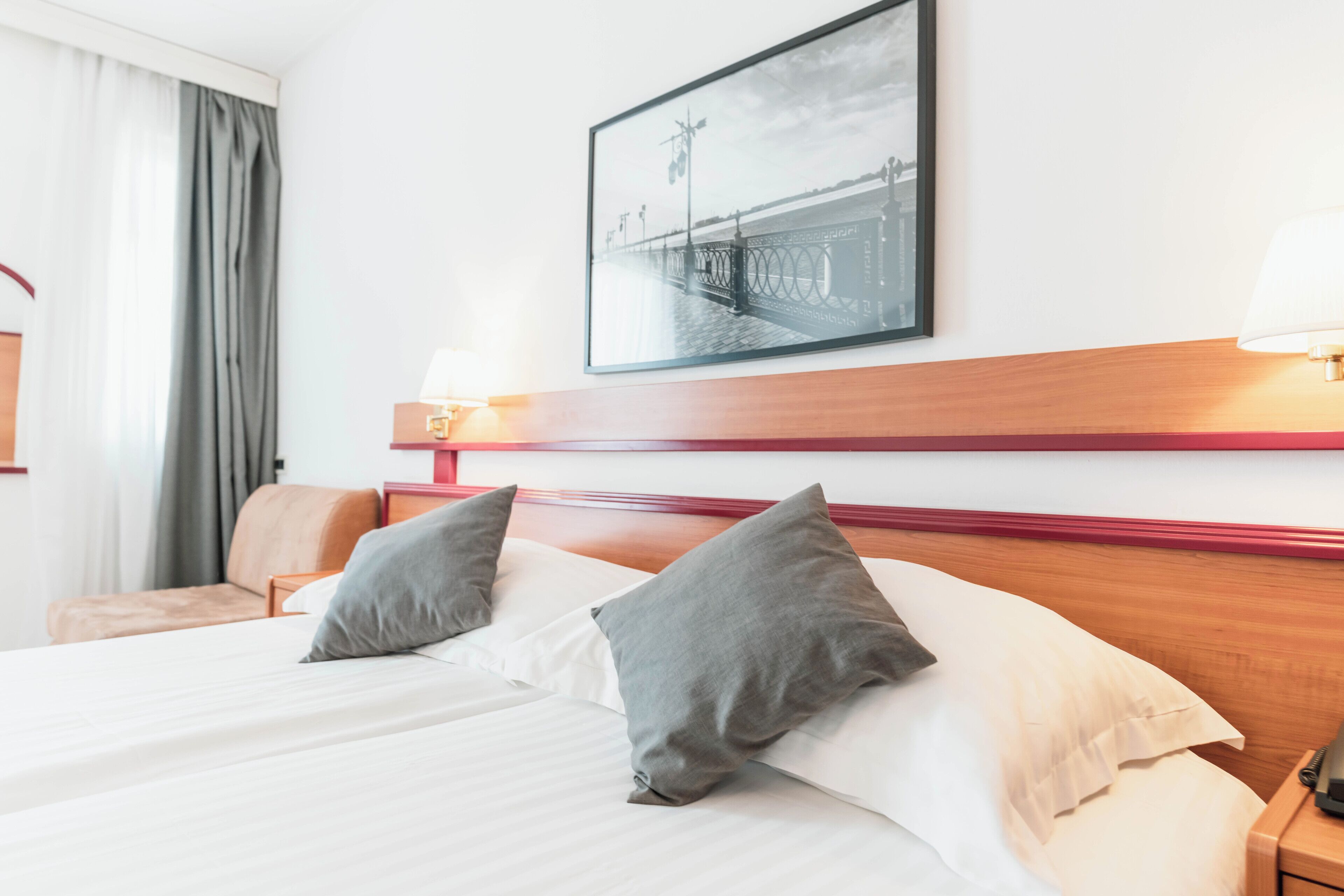 A hotel room with a bed, pillows, a nightstand, a lamp, a framed picture on the wall, and a window with curtains.