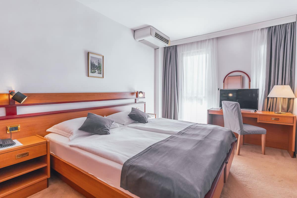 A hotel room with a double bed, a desk with a computer, a chair, a TV, and a bedside table with a lamp.