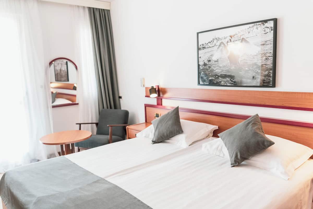 A hotel room with a bed, a chair, a small table, a mirror, and a wall-mounted picture.
