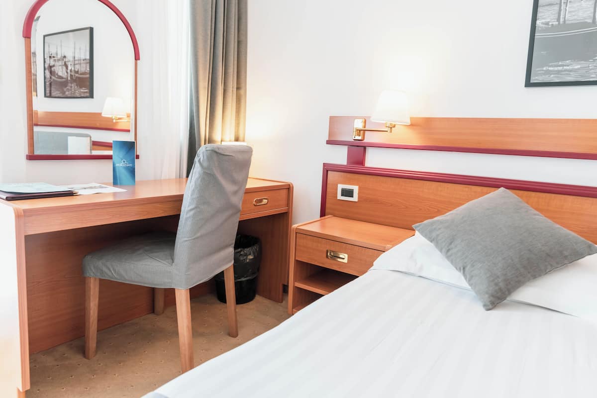 A hotel room with a wooden desk, a chair, a bed with a pillow, a nightstand, a lamp, a trash bin, and a framed picture on the wall.