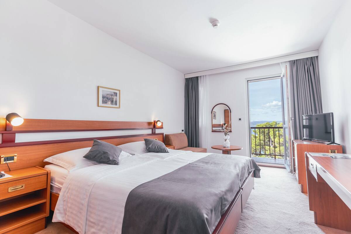 A hotel room with a bed, bedside table, lamp, TV, and a balcony with a view.