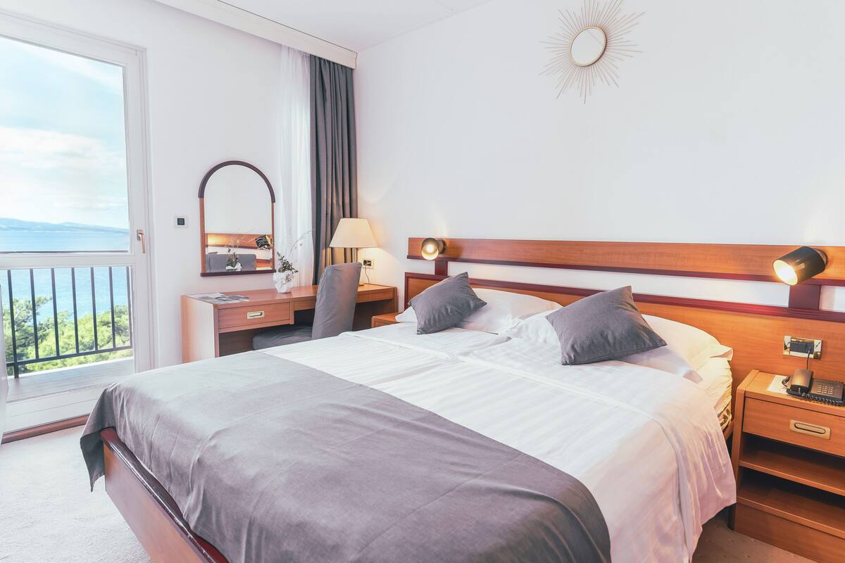 A hotel room with a large bed, a desk with a lamp, a nightstand, and a view of the sea through a balcony.