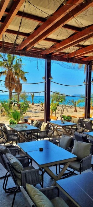 An outdoor seating area with tables and chairs, overlooking a beach with palm trees and the sea. - Hotel San Antonio (Podstrana)