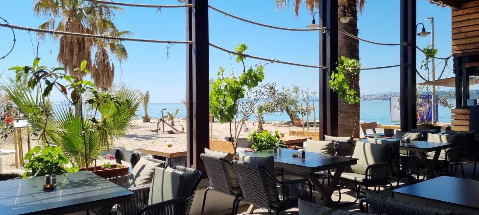An outdoor seating area with tables and chairs, overlooking a beach and the sea.