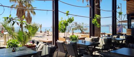 An outdoor seating area with tables and chairs, overlooking a beach and the sea.