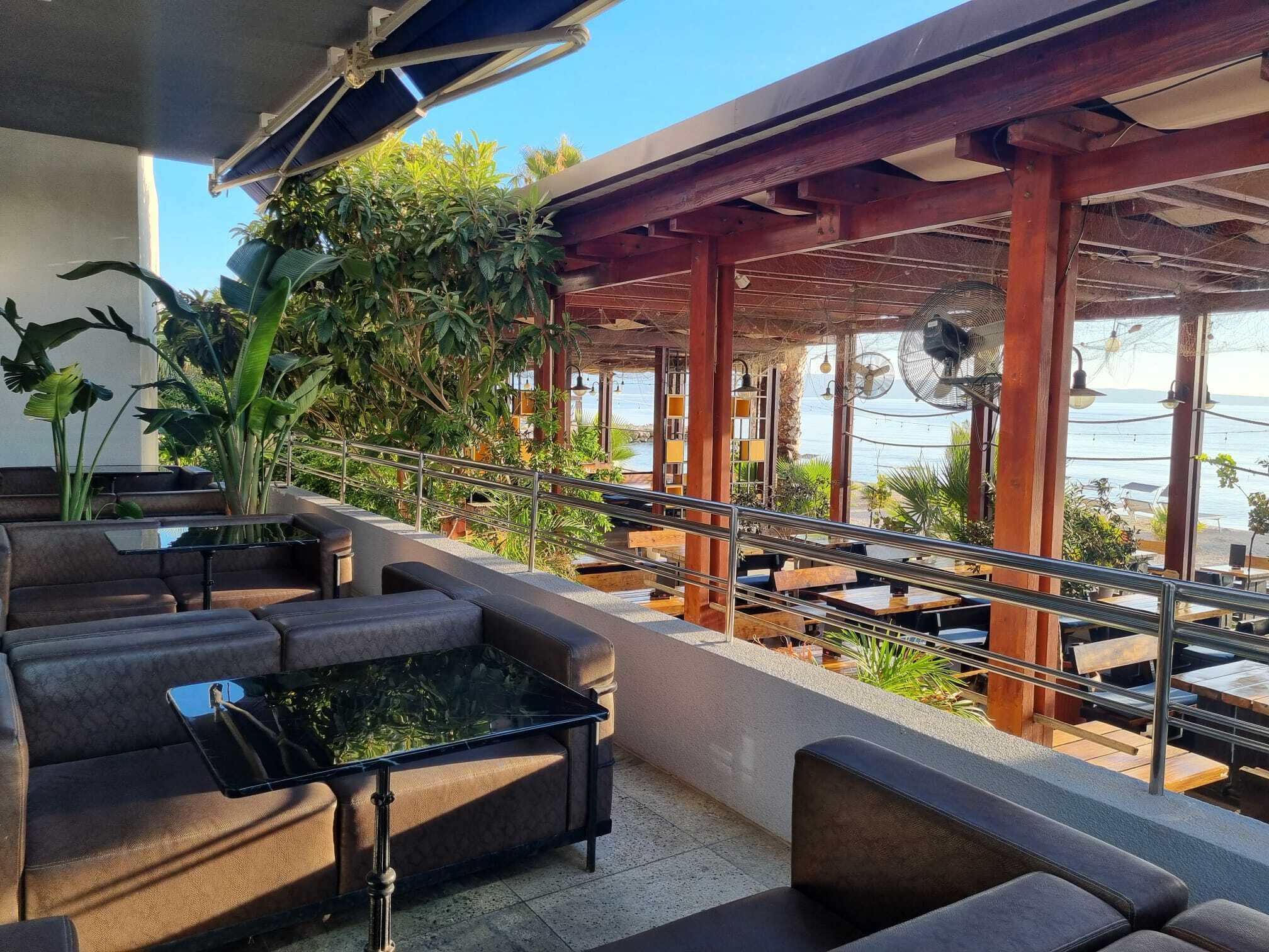 An outdoor seating area with cushioned benches and tables, surrounded by plants and overlooking a body of water.