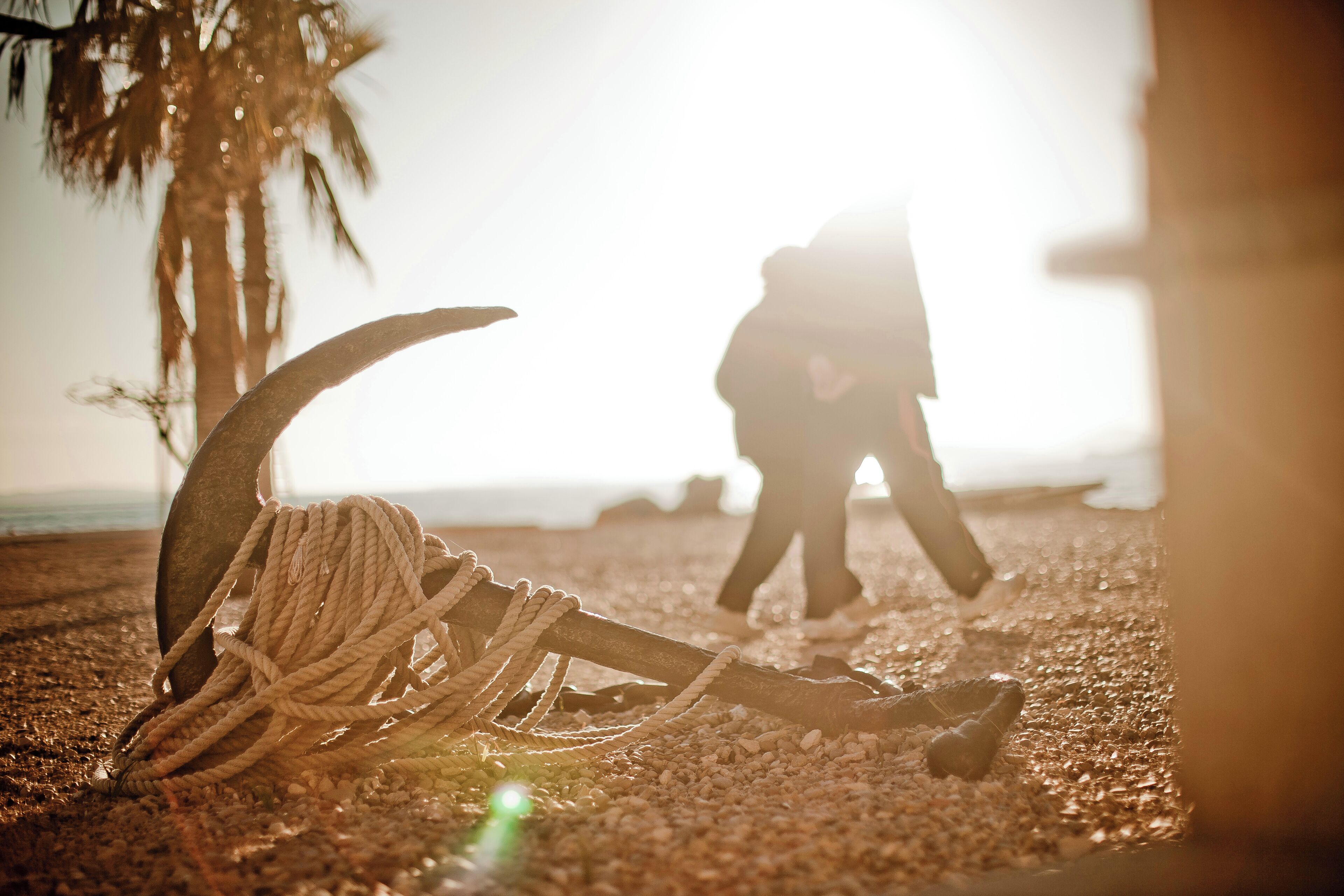 A rope tied to a metal anchor on a pebbled beach with a palm tree and a person in the background.
