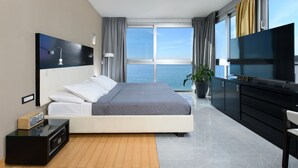 A modern hotel room with a large bed, a TV, a desk, and a view of the sea.