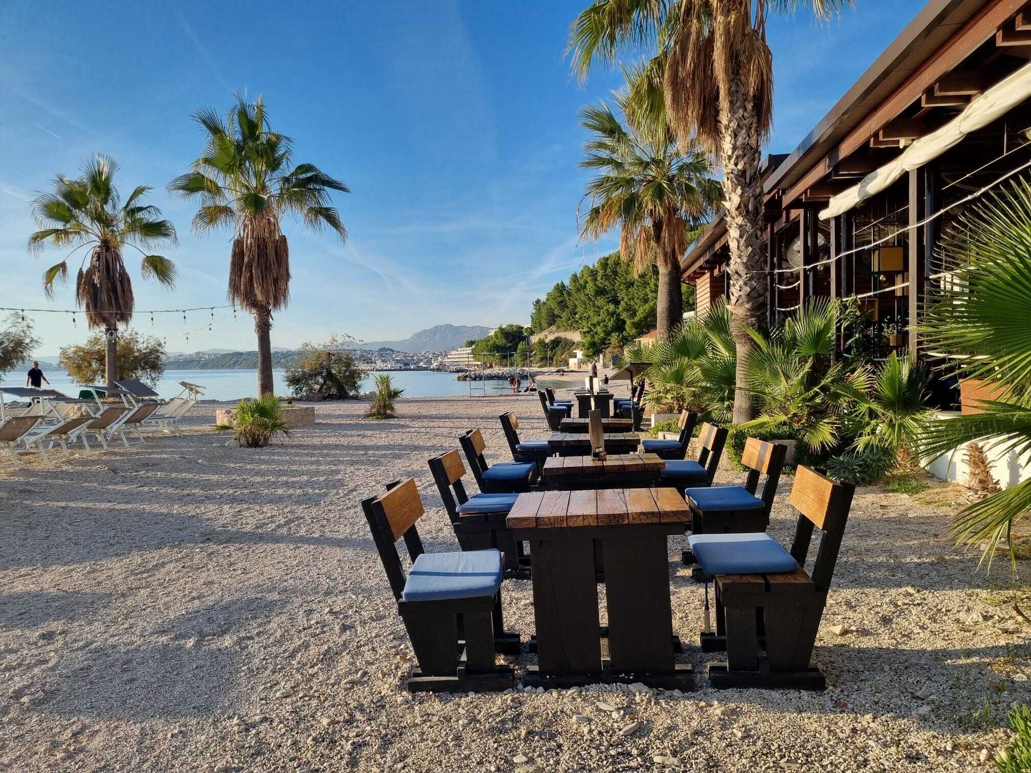 Ein Außenbereich mit Tischen, Stühlen, Palmen und Blick auf das Meer.