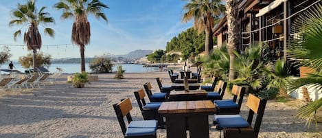An outdoor seating area with tables and chairs, palm trees, and a view of the sea.