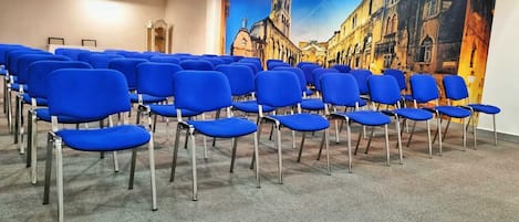Un salón de conferencias con filas de sillas azules, suelo alfombrado y un mural de un paisaje urbano en la pared.