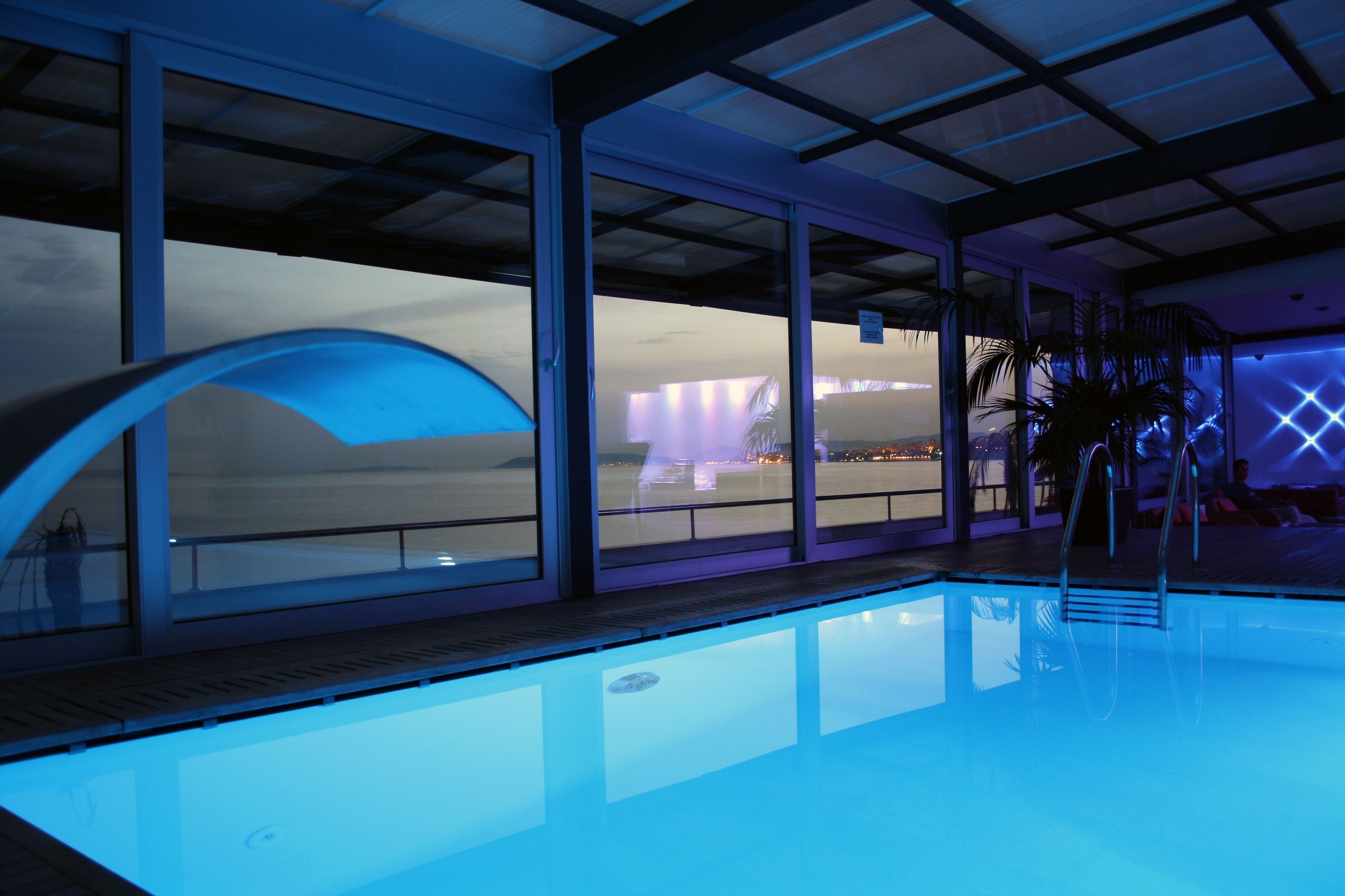 Una piscina interior moderna con paredes de vidrio transparente, una terraza de madera y vistas a la ciudad al anochecer.