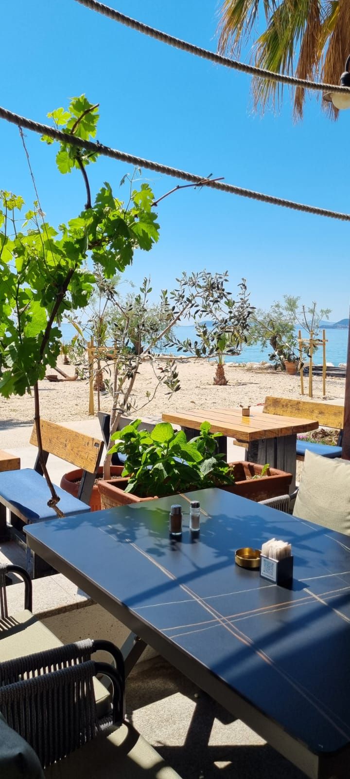 Een eetgedeelte aan het strand met een tafel, stoelen en uitzicht op het strand, plus wat groen.