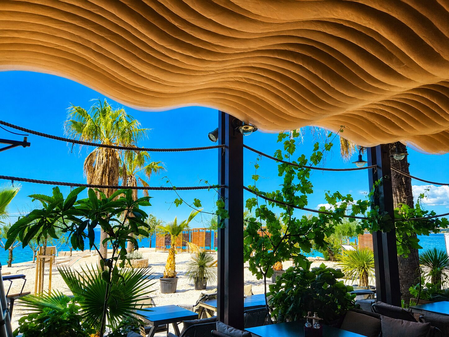 An outdoor seating area with tables and chairs, surrounded by plants and a view of the sea.